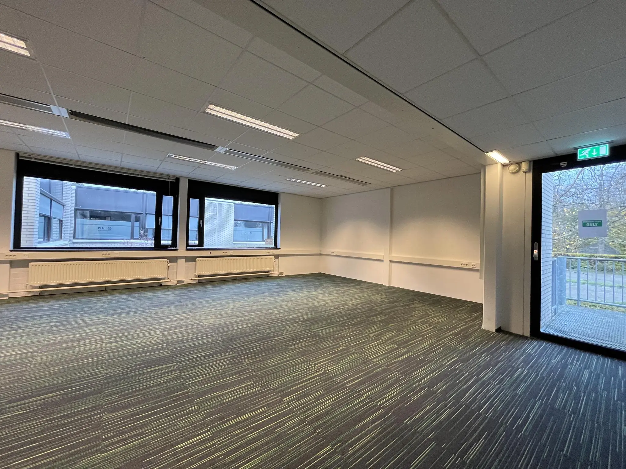 Lege kantoorruimte met tapijtvloer, grote ramen en een nooduitgang aan de Burgemeester Verderlaan.